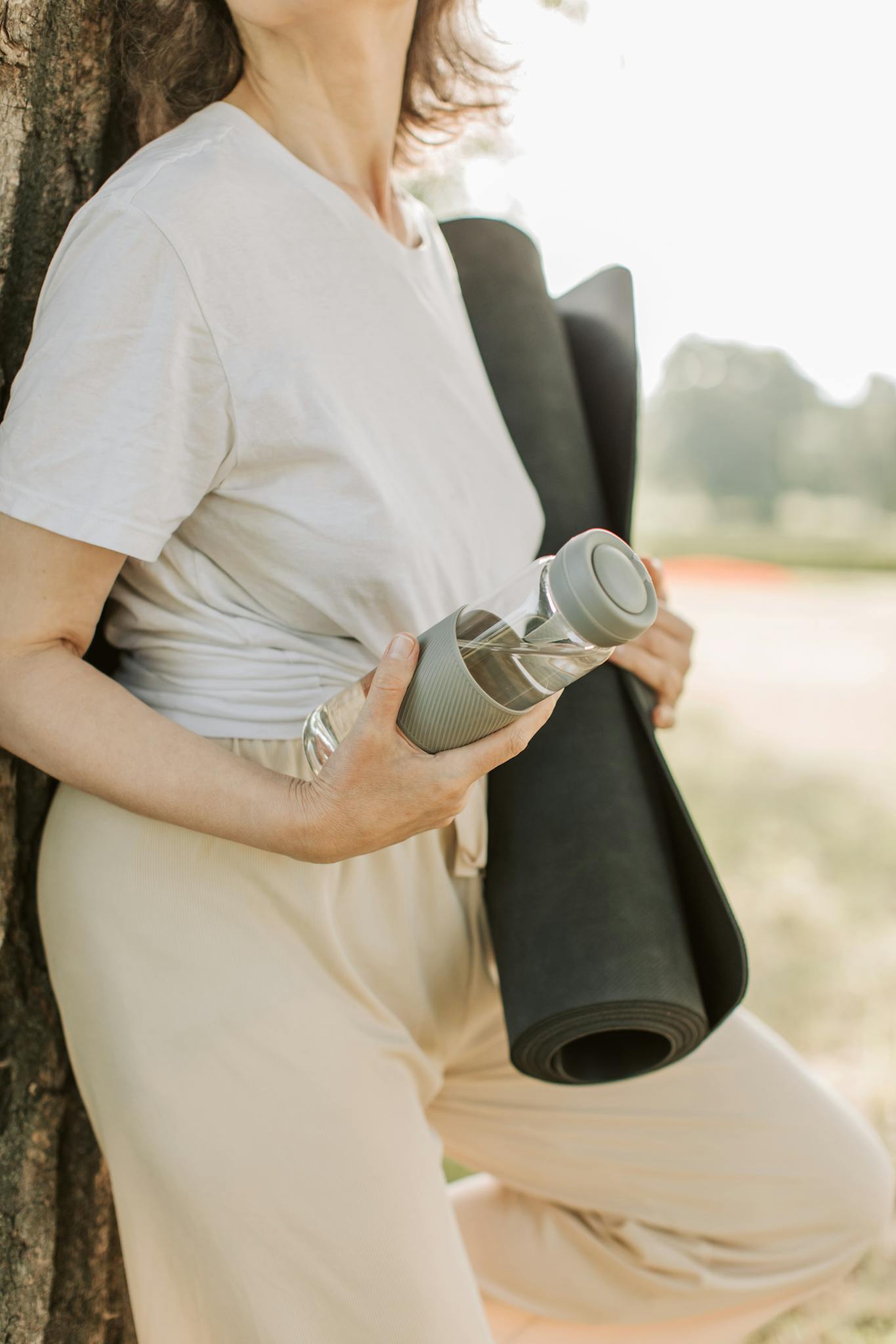 A woman in a white shirt leans on a tree outdoors, holding a yoga mat and water bottle, embracing wellness.
