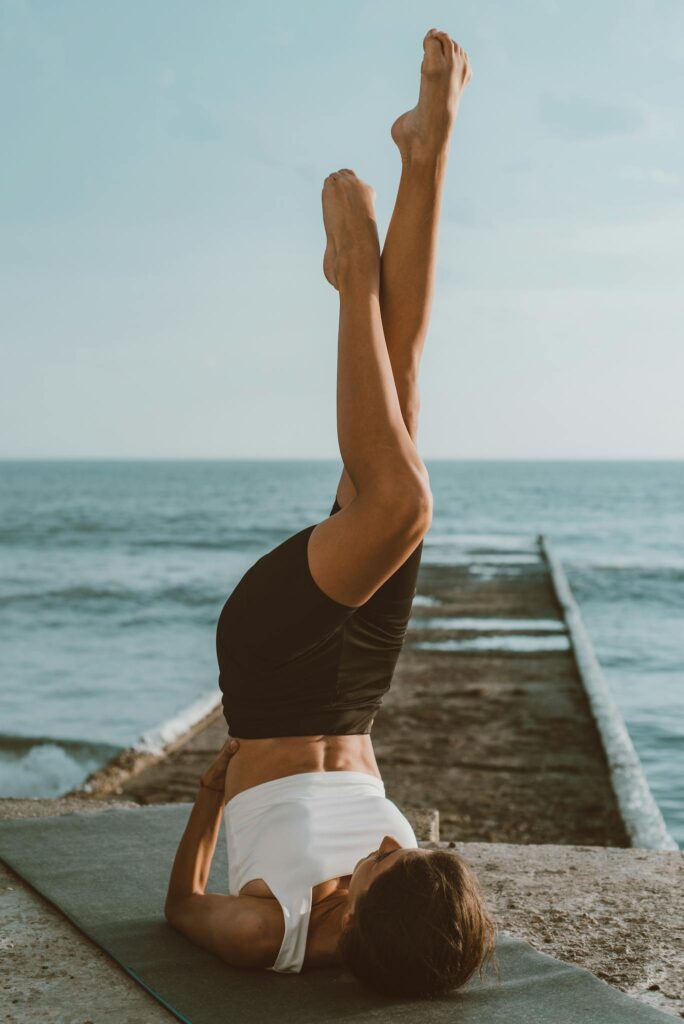 A woman in sports attire performs yoga on a mat by the ocean, emphasizing balance and flexibility.