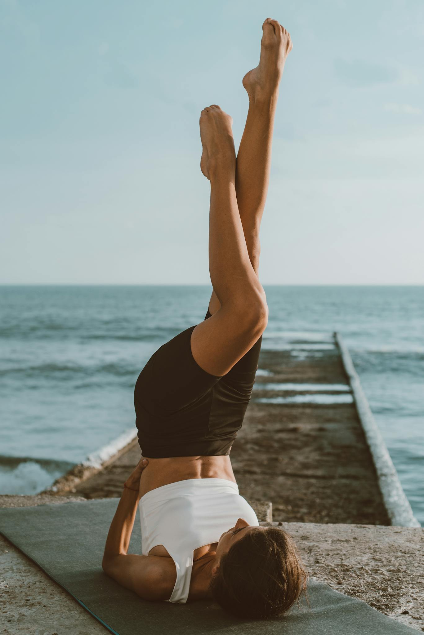 A woman in sports attire performs yoga on a mat by the ocean, emphasizing balance and flexibility.
